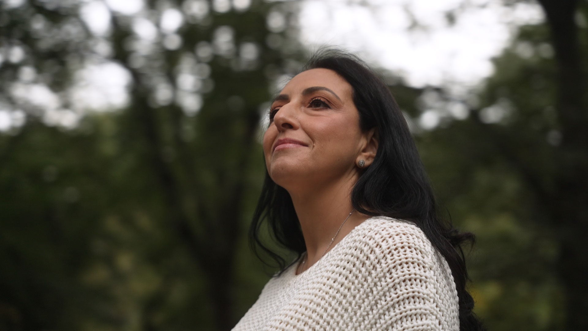 Person looking into the distance while outdoors near trees and buildings.