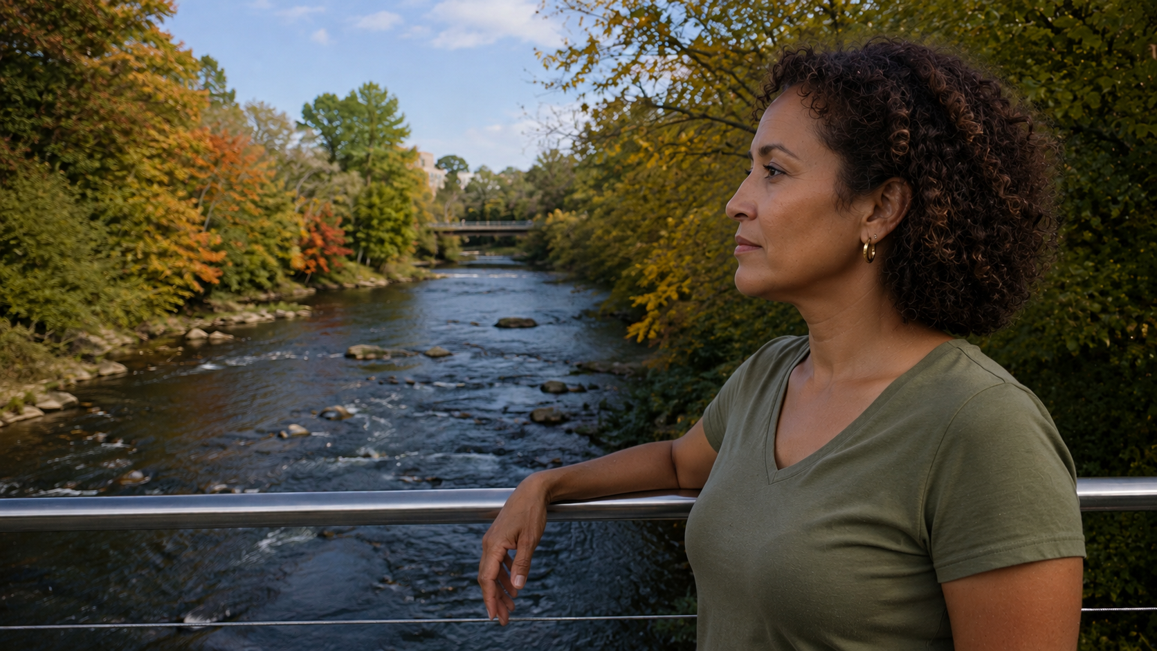 Person standing on a bridge overlooking a tree-lined river with autumn foliage.